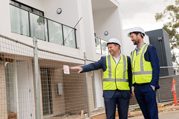 Two men in blue business suits and high visibility vests and white hard hats stand in front of a fenced off, newly developed property and point at vacant space.